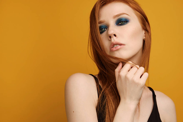 Red-haired girl in a black top and with blue eye makeup stands on a yellow background and looks in the frame, playing with hair - Photo, image