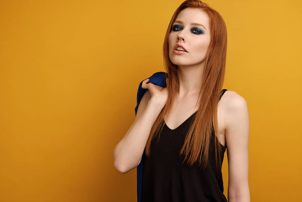 Red-haired girl in a black top and blue smokey stands on a yellow background, throwing his jacket over his shoulder  - Fotografie, Obrázek