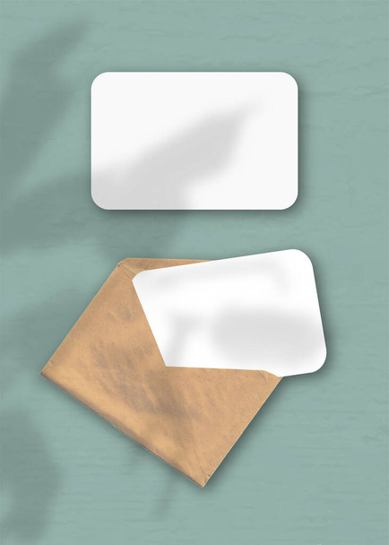 Ein Umschlag mit zwei Blättern strukturiertem weißem Papier auf grünem Hintergrund. Mockup-Overlay mit den Schatten der Pflanze. Natürliches Licht wirft Schatten von einem Zygokaktus. Vertikale Orientierung. - Foto, Bild