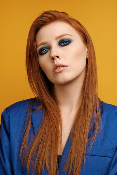Headshot. Redhead girl in a blue jacket and blue smokey stands on a yellow background and looks into the frame with head bowed - Valokuva, kuva