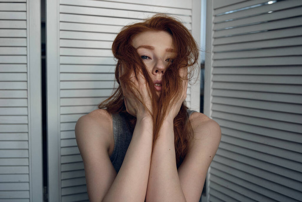 A red-haired girl in a t-shirt and pants sits against a white screen and looks at the camera, covering her face with hair. - Foto, Imagen