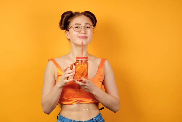 Red-haired girl in round glasses and an orange top is standing on a yellow background, smiling pretty with a lemonade in hands - Foto, Bild