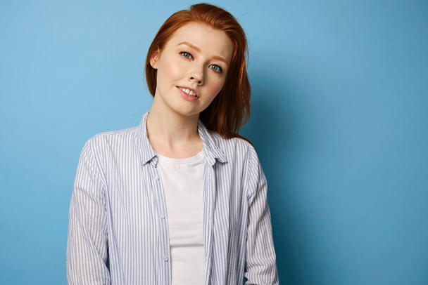 Red-haired girl in a striped shirt and a white T-shirt stands on a blue background with a slight smile and bowing head to the side - Photo, Image