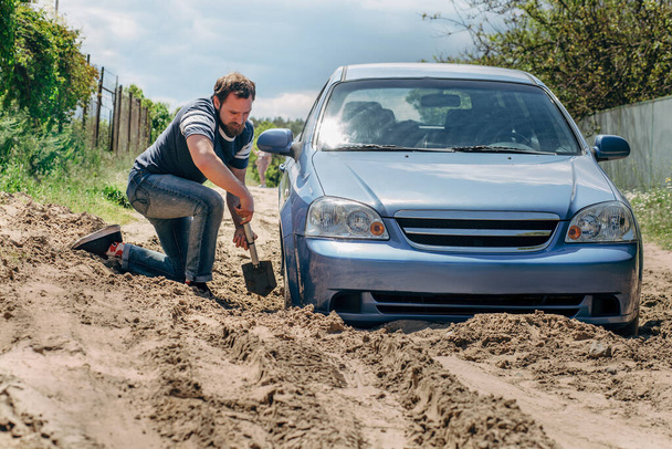 A blue car stuck in the sand.. A strong man with a beard digs a shovel of the wheel of the car.  - Φωτογραφία, εικόνα