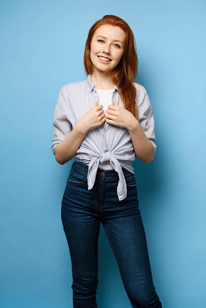 Red-haired girl in a striped shirt tied at the waist stands on a blue background and smiles, holding her hands - Foto, afbeelding
