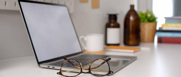 Vue latérale du bureau à domicile avec ordinateur portable maquette, lunettes, tasse et décorations sur table blanche
  - Photo, image