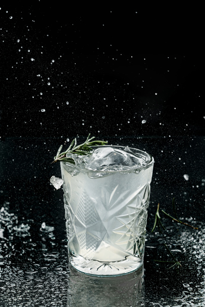 Свежий алкогольный коктейль с водкой, лед, розмарин в стакане на черном фоне. Студия выпивки в замерзающем движении, летающий лёд и капли. Летний холодный напиток и коктейль
 - Фото, изображение