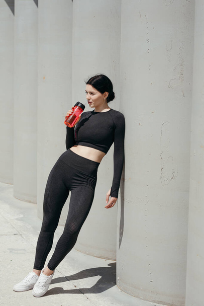 Pilates Asian trainer leans on concrete columns on the embankment and drinks water from a bottle. - Photo, Image