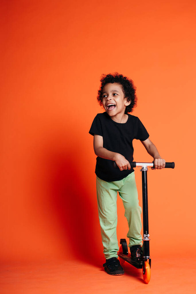 Full-length portrait of an Ethiopian boy laughing and standing on a scooter on an orange background in the studio - Fotó, kép