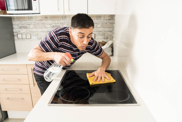 Man in casual clothes cleaning the induction hob in the kitchen with a cloth. Mid shot. - 写真・画像