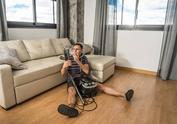 Man sitting on the floor in the living room trying to understand how a vacuum cleaner works. Full body. - Photo, image