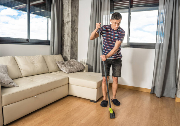 Man in casual clothes and slippers sweeping his living room. Full body. - Foto, afbeelding