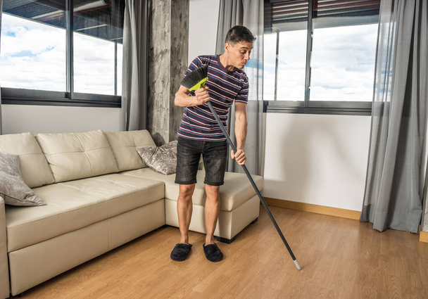 Uomo in abiti casual e pantofole cercando di capire come funziona una scopa nel suo soggiorno. Corpo completo
. - Foto, immagini