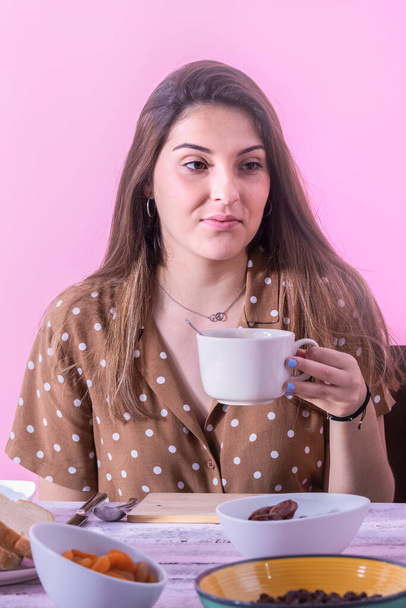 Young beautiful woman holding a cup with an assortment of bowls with dry fruit on a table on a pink background. Vegan breakfast and healthy food concept. - Fotó, kép
