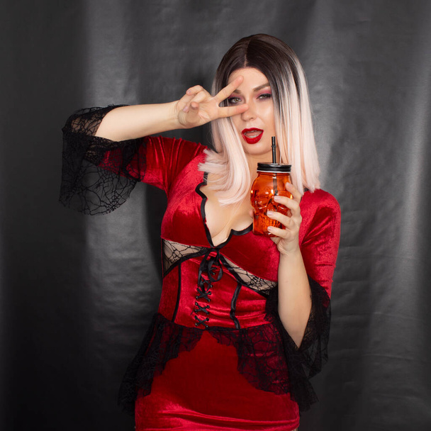 Young beautiful blonde woman in a red vampire dress holds an orange glass in the shape of a skull in her hands - Foto, Imagem