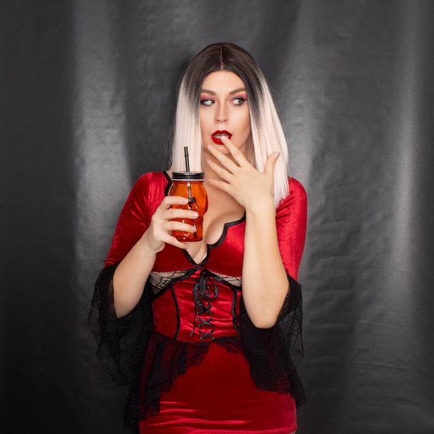 Young beautiful blonde woman in a red vampire dress holds an orange glass in the shape of a skull in her hands - Foto, afbeelding