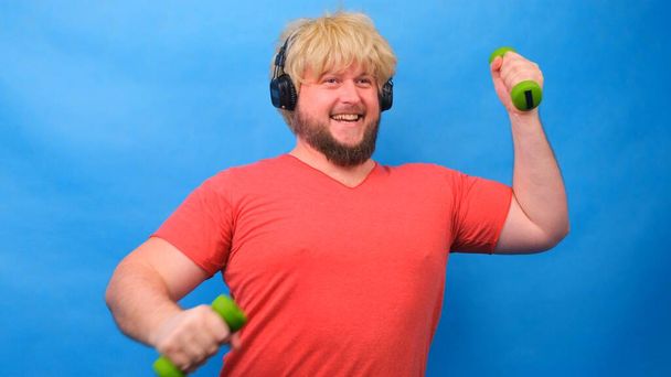Engraçado homem gordo esquisito em uma peruca e uma camiseta rosa com halteres fazendo exercícios em um fundo azul
. - Foto, Imagem