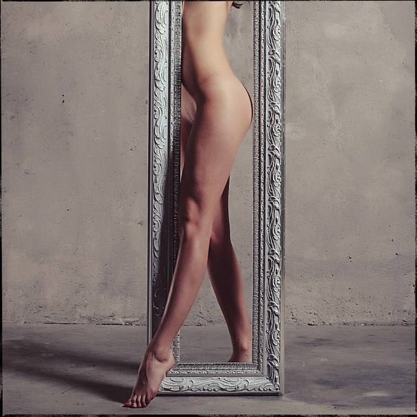 Young beautiful slender girl, naked in the studio against a gray wall, she holds in her hands an empty frame from the mirror - Foto, Bild
