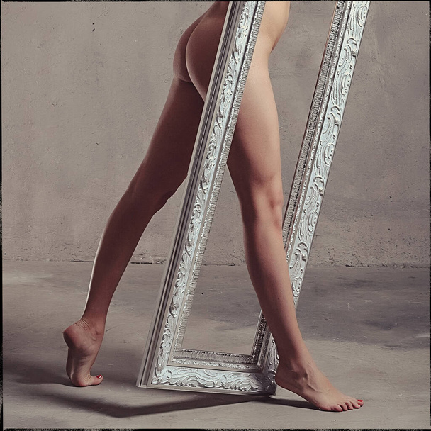 Young beautiful slender girl, naked in the studio against a gray wall, she holds in her hands an empty frame from the mirror - Φωτογραφία, εικόνα