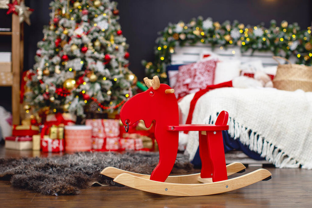 happy holiday season! Christmas decoration, interior decorated for the holiday, with an elegant Christmas tree, a bed and a large number of gifts. In the foreground is a wooden toy rocking moose. - Photo, Image