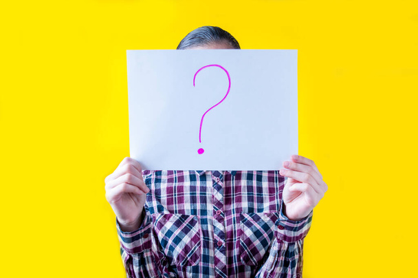 Girl in a plaid shirt on a yellow background holding a white sheet of paper with a drawn question mark - Foto, immagini