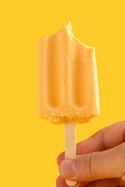 hand holding mango pudding flavor popsicle with couple of bites on a yellow background - Zdjęcie, obraz