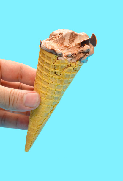 χέρι κρατώντας μια φρέσκια γεύση σοκολάτας παγωτό χωνάκι με μερικά τσιμπήματα σε μπλε φόντο - Φωτογραφία, εικόνα