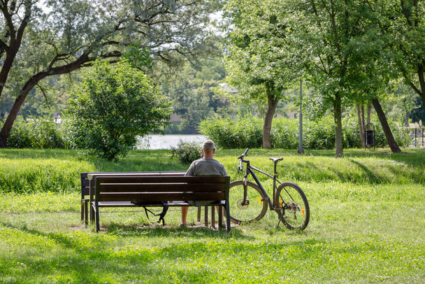 自転車に乗って湖のそばのベンチに腰を下ろしている高齢者スポーツ灰色の髪の男。美しい自然景観。高齢者コンセプトでのアクティブなライフスタイルとスポーツ活動 - 写真・画像