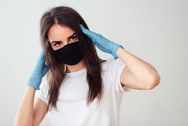 Portrait of a girl in a black medical mask and a white T-shirt on a white background, hands in gloves near the head. Symptoms of the common cold. Stay home concept - Photo, Image