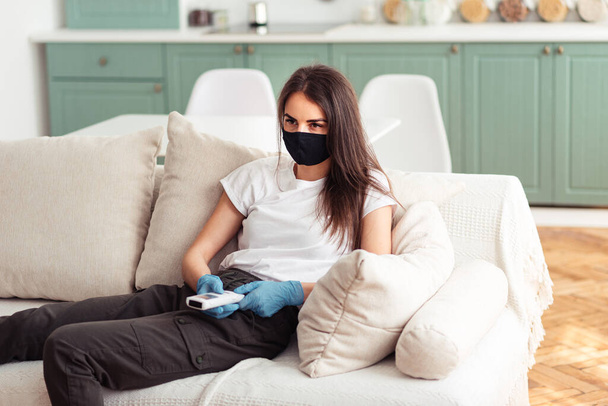 egy hideg és szomorú lány maszkban és kesztyűben ül a kanapén és egy távirányítót tart a kezében. Az otthon maradás fogalma. - Fotó, kép