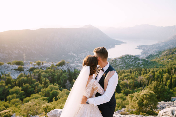 Svatební pár stojí na vrcholu hory s panoramatickým výhledem na záliv Kotor, při západu slunce. Ženich jemně objímá nevěstu. Svatební fotografie výtvarného umění v Černé Hoře, Mount Lovchen. - Fotografie, Obrázek