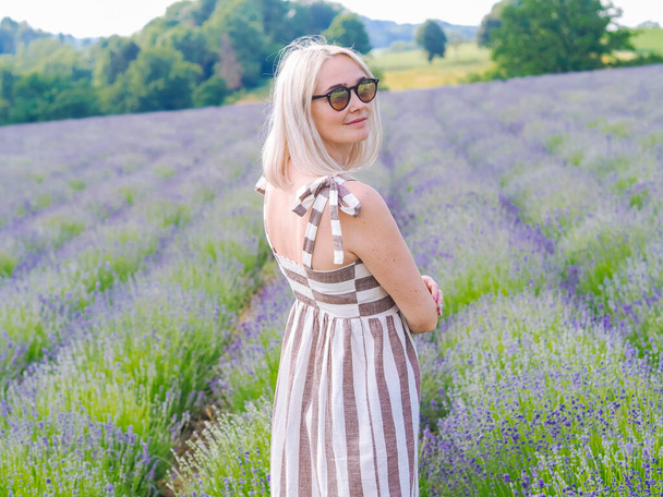 Beautiful blond woman in dress sit between lavender fields in Provence. Violet lavender fields blooming in summer sunlight. Sea of Lilac Flowers landscape. Bunch of scented flowers of French Provence. - Photo, Image