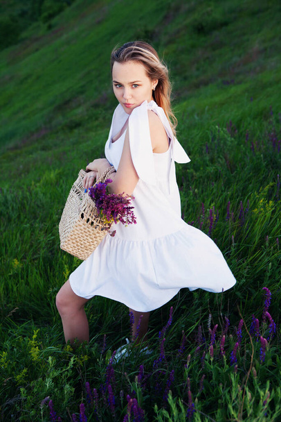 Hermosa chica en vestido blanco con bolsa de paja y flores silvestres en el prado
. - Foto, Imagen