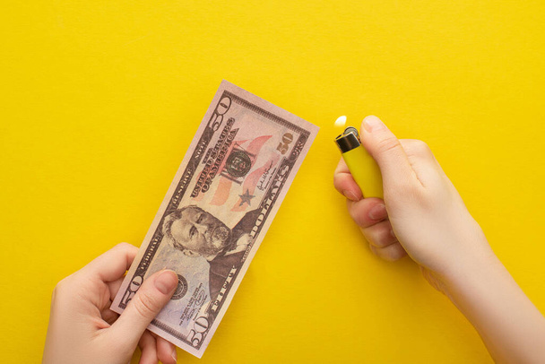 KYIV, UKRAINE - MARCH 25, 2020: cropped view of woman holding lighter near dollar banknote isolated on yellow  - Foto, Bild