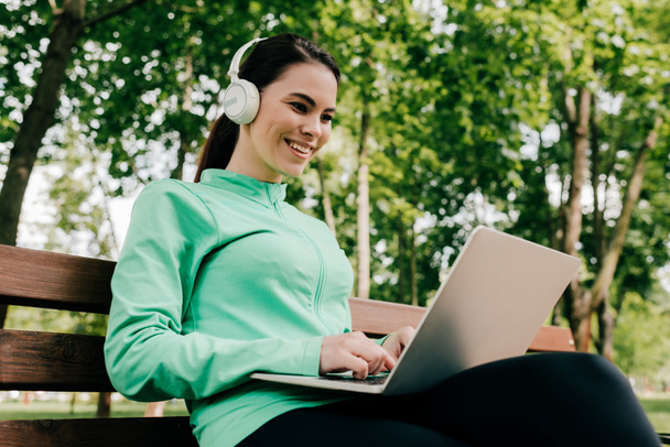 Smiling girl in headphones using laptop on bench in park  - Photo, Image