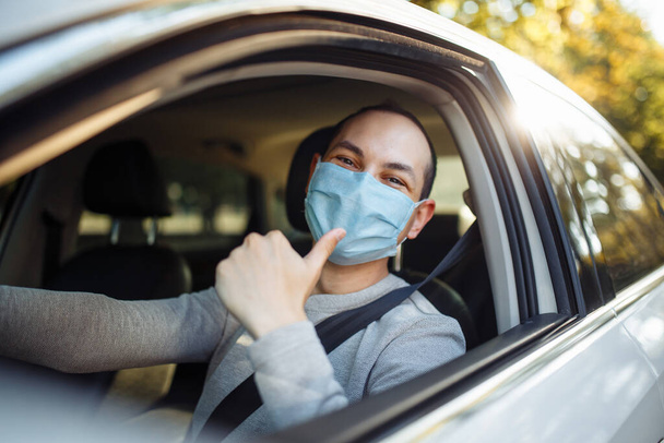 Young man shows thumb up like sign wearing sterile mask in the car. Taxi driver delivers his passanger to the destination. Social distance, new normal, virus spread prevention and treat concept - Fotoğraf, Görsel