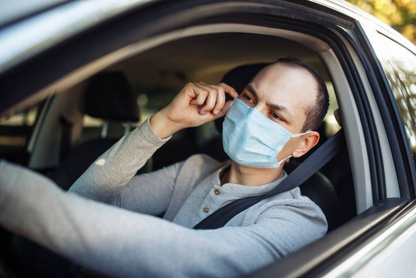 Een man rijdt in een auto en past een medisch masker aan tijdens een coronavirus uitbraak. Taxi chauffeur brengt zijn passagier naar de bestemming. Sociale afstand, nieuw normaal, virusverspreidings- en behandelingsconcept - Foto, afbeelding