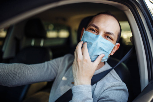 Un hombre conduce un coche y ajusta la máscara médica durante el brote de coronavirus. El taxista lleva a su pasajero al destino. Distancia social, nueva normalidad, prevención y tratamiento de la propagación del virus
 - Foto, imagen
