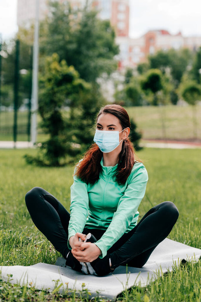 Selective focus of young sportswoman in medical mask warming up on fitness mat in park  - Foto, Imagem