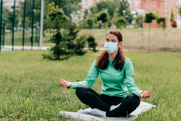 Young woman in medical mask meditating on fitness mat in park  - Foto, imagen