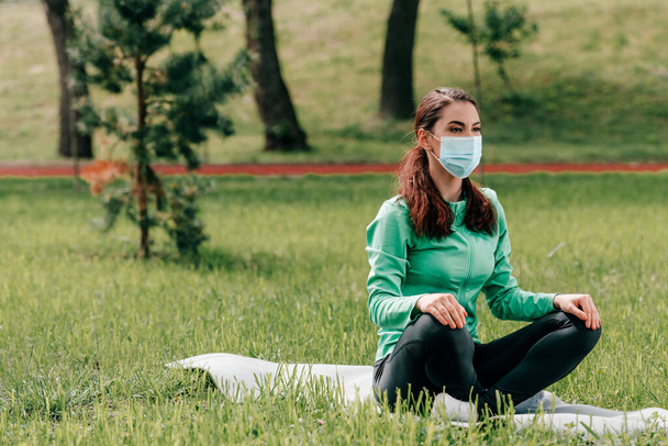 Kobieta w masce medycznej siedzi na macie fitness w parku  - Zdjęcie, obraz
