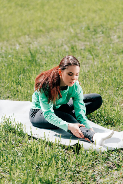Selective focus of attractive sportswoman stretching on fitness mat on grass in park  - Photo, Image