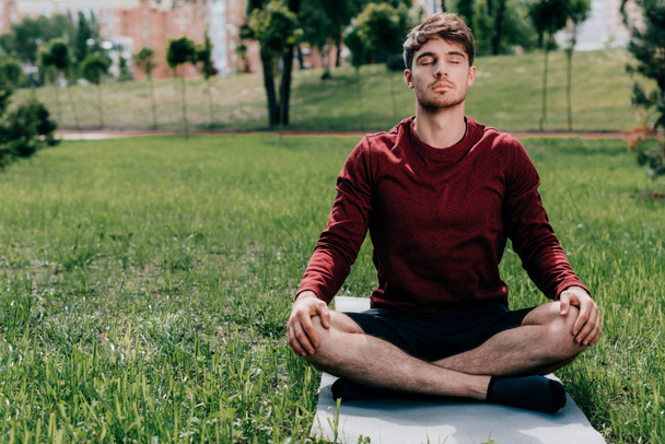 Yakışıklı genç adam parkta fitness minderinde otururken meditasyon yapıyor.  - Fotoğraf, Görsel