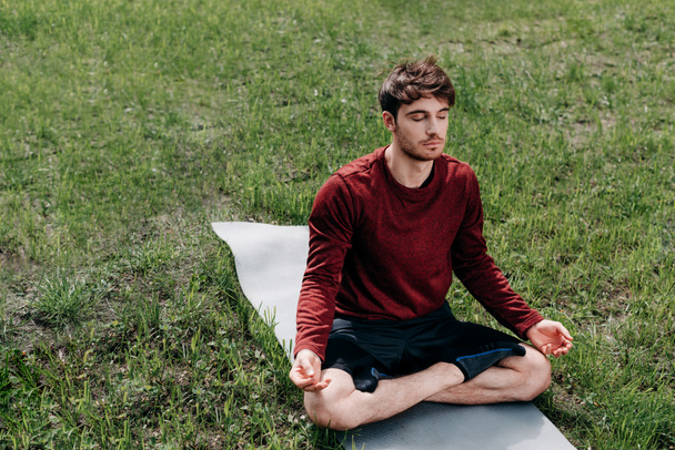 Jovem meditando no tapete de fitness na grama no parque
  - Foto, Imagem