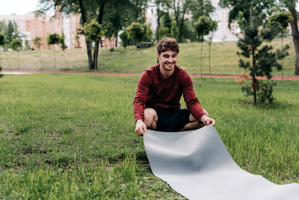Jóképű férfi mosolyog, miközben fitneszszőnyeget terít a fűre a parkban.  - Fotó, kép