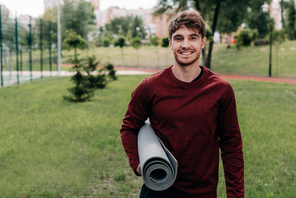 Handsome sportsman smiling at camera while holding fitness mat in park  - Fotó, kép