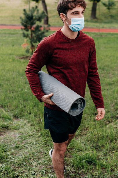 Αθλητής με ιατρική μάσκα κρατώντας χαλάκι γυμναστικής ενώ περπατούσε στο πάρκο  - Φωτογραφία, εικόνα