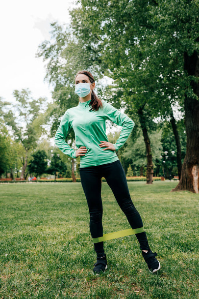 Sportswoman in medical mask working out with resistance band in park  - Foto, imagen