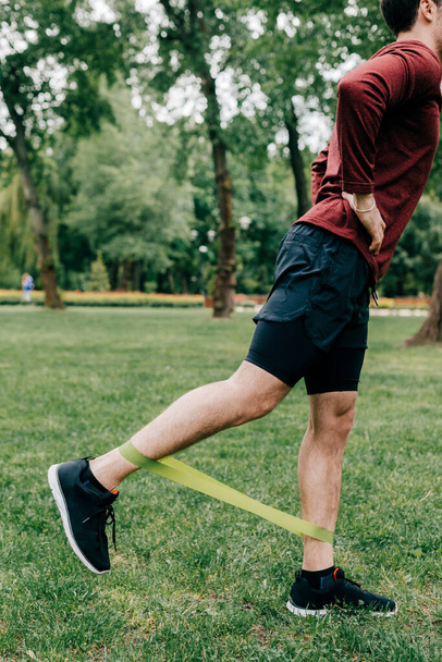 Cropped view of sportsman using elastics band while training on lawn in park  - Foto, Imagem