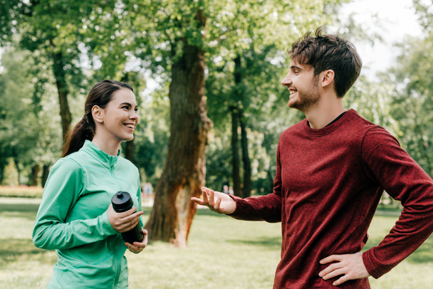 Deportista sonriente hablando con novia con botella deportiva en el parque
  - Foto, Imagen